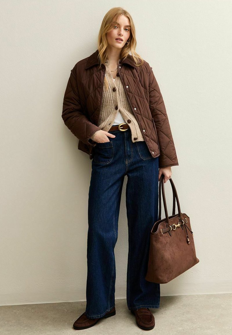 Veste matelassée marron sur un pull en maille beige, jeans larges bleus, sac à main marron foncé et chaussures assorties. Simples accents métalliques sur le sac.