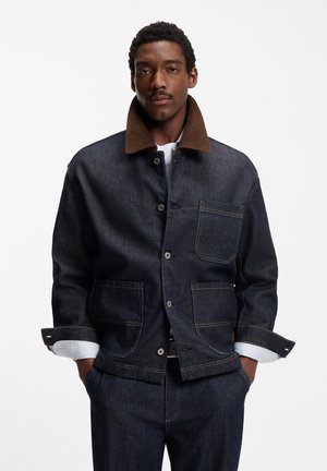 Mand iført mørk denimjakke med brun krave og matchende mørke denimbukser, stående med hænderne i lommerne mod en hvid baggrund.