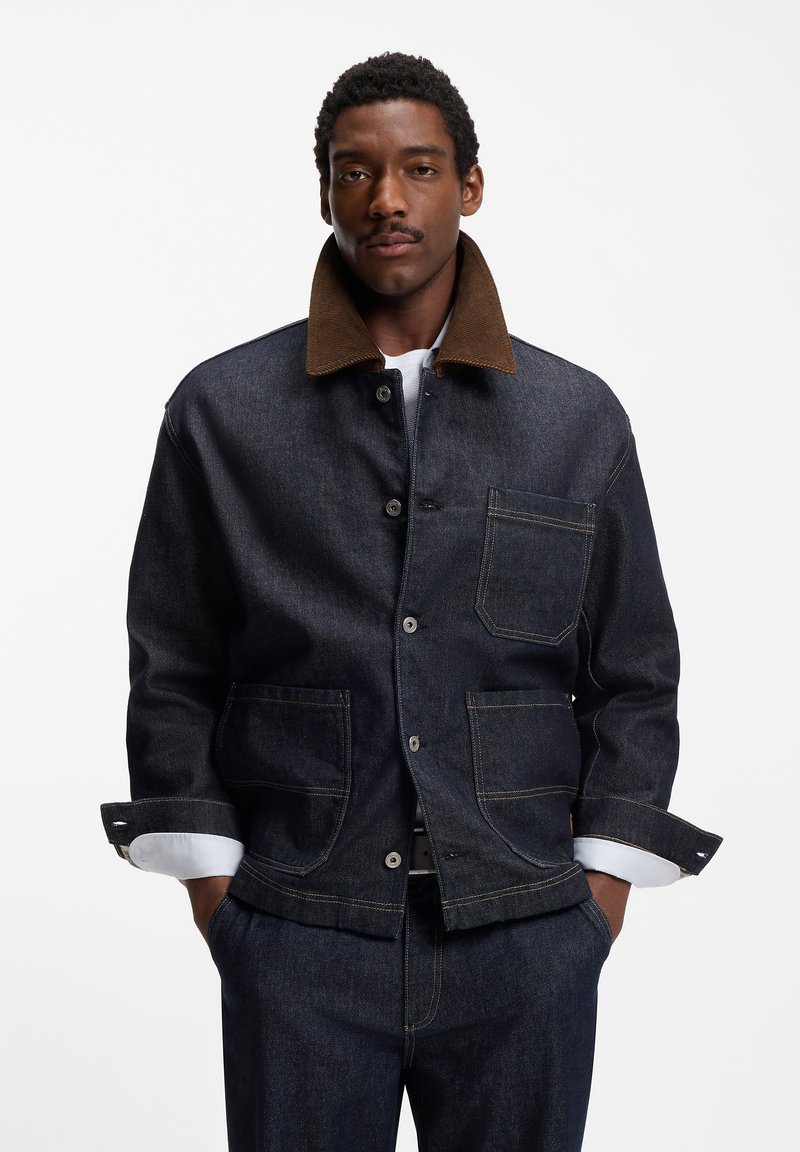 Man wearing dark denim jacket with brown collar and matching dark denim pants, standing with hands in pockets against white background.