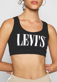 Svart croptop i bomull med vid halsringning och stort vitt "LEVI'S"-logotyp på framsidan. Slät textur med en figurnära passform.