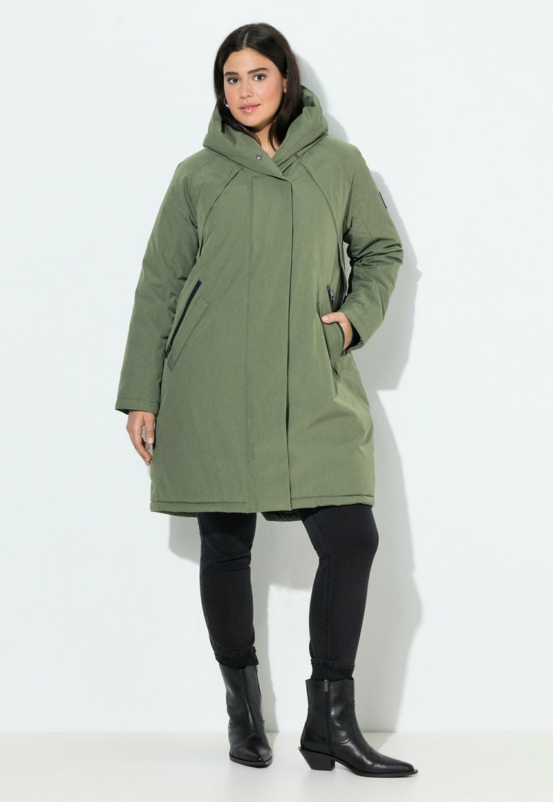 Cappotto lungo verde oliva con cappuccio, design strutturato, tasche laterali con zip e texture liscia, abbinato a pantaloni neri aderenti e stivali alla caviglia.