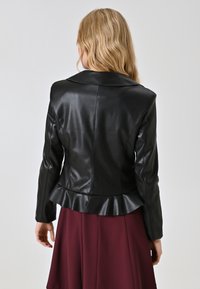 Giacca nera in similpelle con orlo a peplum, vestibilità aderente e collo ampio. Modello con indosso una gonna bordeaux, vista da dietro.