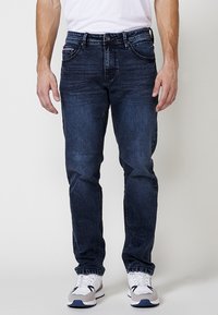 Jeans bărbați de culoare albastru închis, din denim, cu croi regulat, design clasic cu cinci buzunare și decolorare subtilă. Purtați cu adidași albi.