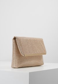 Béžová kabelka na ruku s texturovaným látkovým povrchem, se zlatým kovovým akcentem na přední straně a zavíráním na klopu.