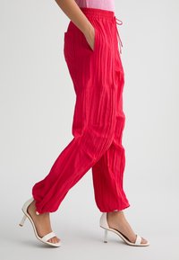 Pantaloni rossi leggeri, raggrinziti, con cordoncino, tasche laterali, caviglie affusolate e un fit rilassato, abbinati a sandali bianchi con tacco.