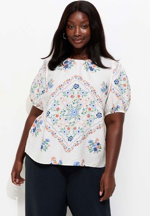 Vrouw met lang donker haar, gekleed in een witte blouse met pofmouwen en kleurrijke bloemmotieven, gecombineerd met een zwarte broek, staand tegen een witte achtergrond.