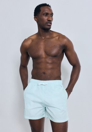 Μυώδης άνδρας με κοντά μαλλιά, ντυμένος με ανοιχτό μπλε σορτς κολύμβησης, στέκεται με τα χέρια στις τσέπες, κοιτώντας ελαφρώς προς το πλάι σε απλό φόντο.