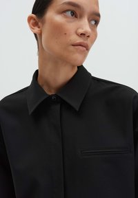 Vrouw met kort haar in een zwarte overhemd met kraag, kijkend naar de zijkant tegen een effen lichte achtergrond.