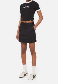 Camiseta corta negra con ribete blanco que presenta el logo "DICKIES", combinada con una falda negra. Usada con calcetines blancos y zapatillas.