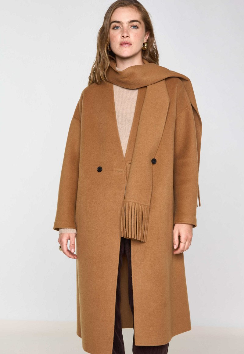 Cappotto di lana marrone con ampio rever, chiusura doppiopetto e due bottoni neri. Presenta una lunga sciarpa per il collo con frange.