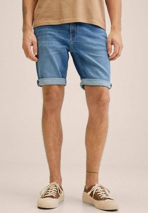 Mann trägt hellblaue, hochgekrempelte Jeansshorts, beigefarbene Sneakers mit weißen Schnürsenkeln und ein beiges Hemd, steht mit entspannt an den Seiten hängenden Händen.