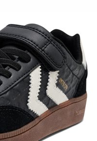 Sneaker nero con una combinazione di pelle strutturata e camoscio, con accenti bianchi a zigzag, una fascia in Velcro e una suola in gomma color gum.