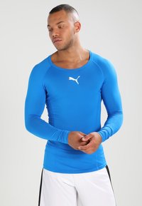 Maglietta sportiva a maniche lunghe di colore blu brillante, realizzata in tessuto liscio e elasticizzato. Presenta un collo rotondo e un logo bianco sul petto.