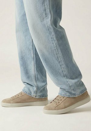 Persoon met lichtblauwe spijkerbroek en beige suède sneakers die op een lichte achtergrond naar rechts kijkt.