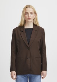 Blazer marron en tissu lisse, avec un col cranté, deux boutons et des poches latérales. Porté sur un haut noir et un jean.
