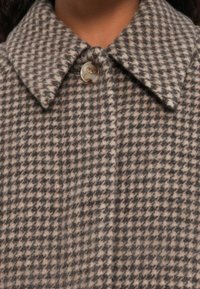 Uldblandingsfrakke med et houndstooth-mønster i grå og cremé, med en stor krave og klar knaplukning. Blød tekstur.