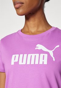 Violetinės medvilnės marškinėliai su balta PUMA logotipu ir šuoliuojančio panteros dizainu. Suapvalinta apykaklė ir trumpomis rankovėmis, lygus paviršius.