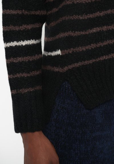 Plano detalle de una mano que lleva un suéter a rayas negro y marrón con una raya blanca, combinado con pantalones oscuros azules texturizados.