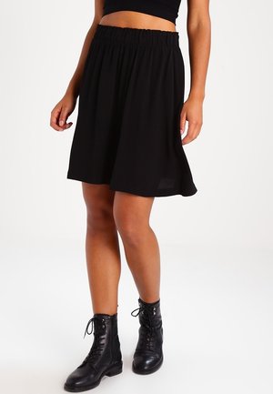 Kvinde iført en sort knælængde nederdel, sort crop top og sorte snørebodystøvler, som står mod en ensfarvet baggrund.