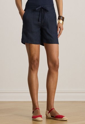Femme portant un short à cordon bleu marine, des espadrilles rouges plates et des bracelets dorés et écaille de tortue, debout sur un sol en bois clair.
