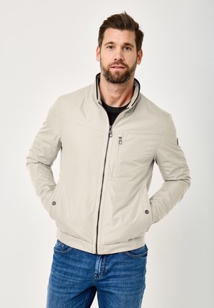Man with short brown hair and beard wears light beige zip-up jacket with hands in pockets and blue jeans, standing against white background.