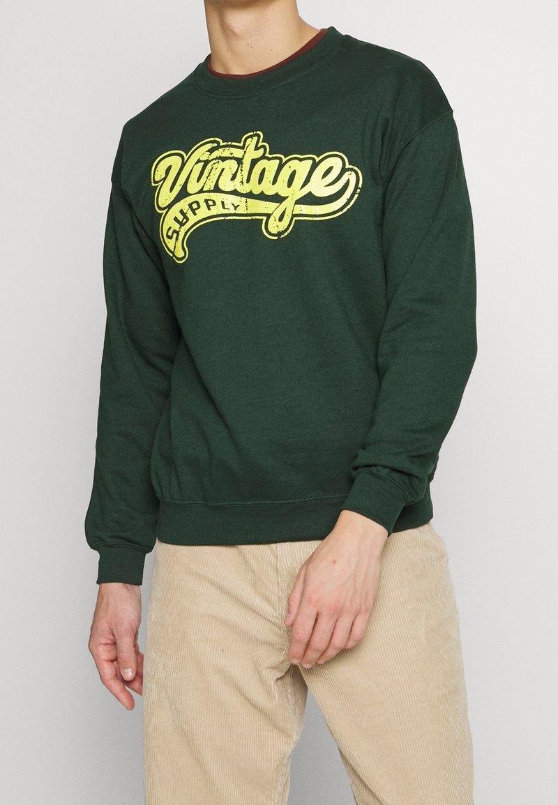 Zelená mikina se žlutým vintage textem "Vintage Supply" na přední straně. Obsahuje kulatý výstřih a manžety s ribbovaným vzorem. Vyrobeno z bavlny.