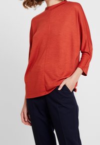 Rotes, leichtes Strickoberteil mit hohem Ausschnitt und dreiviertel Ärmeln, das einen geraden Schnitt und subtile Seitennahtdetails aufweist.