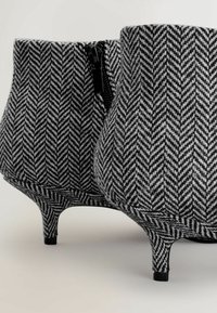 Fekete-fehér hounds-tooth mintás bokacsizma, texturált anyagból, ívelt sziluettel és karcsú tűsarkakkal; oldalt cipzáras záródás jellemzi.