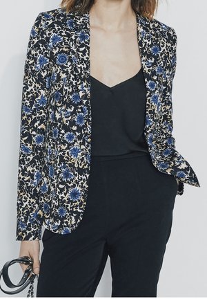 Frau trägt schwarzen floral gemusterten Blazer mit blauen und beigen Blumen, schwarzes ärmelloses Top, schwarze Hose und hält einen schwarzen Taschengurt in der linken Hand.