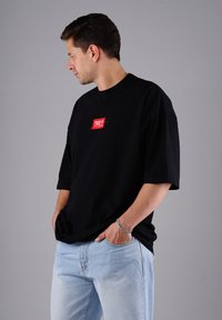 Mann steht mit den Händen in den Taschen, trägt ein schwarzes, übergroßes T-Shirt mit rotem Logo und hellblauen Jeans vor einem schlichten grauen Hintergrund.