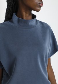 Top cu mânecă scurtă dintr-un material moale, neted, de culoare albastru închis, cu guler înalt și o ușoară strălucire. Decolteu ajustat, cu o formă relaxată a corpului.