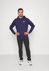 Marineblauwe hoodie met een voorzak, een capuchon met trekkoord en een wit logo. Gecombineerd met zwarte broek en witte sportschoenen.