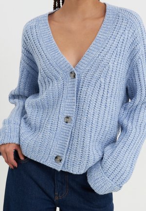 Cardigan bleu clair en maille épaisse avec de gros boutons, col en V profond et manches longues, associé à un jean bleu foncé.