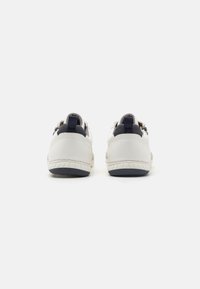 Sneakers din piele albă cu un accent bleumarin la călcâie, talpă din cauciuc texturat și două brâuri pentru o ușurință în încălțare, văzute din spate.