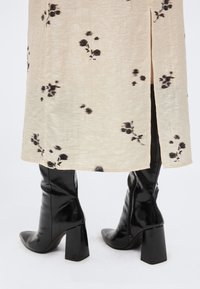 Κρεμ φούστα με μαύρο λουλουδάτο σχέδιο και πλαϊνή σχισμή, φορεμένη με γυαλιστερές μαύρες μπότες γόνατο με ψηλό τακούνι, σε λευκό φόντο.