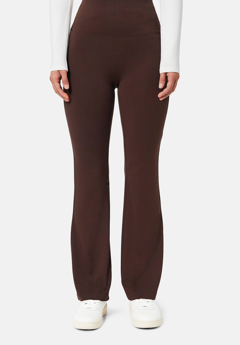 Personne portant un pantalon évasé taille haute marron foncé et des baskets blanches, les mains détendues le long du corps, sur un fond clair uni.