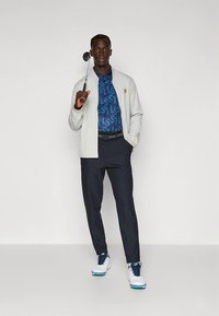 Világosszürke dzseki kék mintás póló fölött, sötétkék nadrág, fehér golfcipő, kezében golfütő. Egyszerű, kényelmes szabású kialakítás.