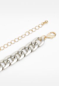 Silberne, grobe Kettenarmband mit glänzender Oberfläche, ausgestattet mit einem goldfarbigen Karabinerverschluss und einem kleinen goldenen Kugelakzent an einem verlängerten Glied.