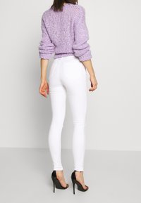 Suéter de punto lila con mangas oversized, combinado con jeans ajustados blancos. El conjunto incluye elegantes zapatos de tacón negro y un diseño minimalista.