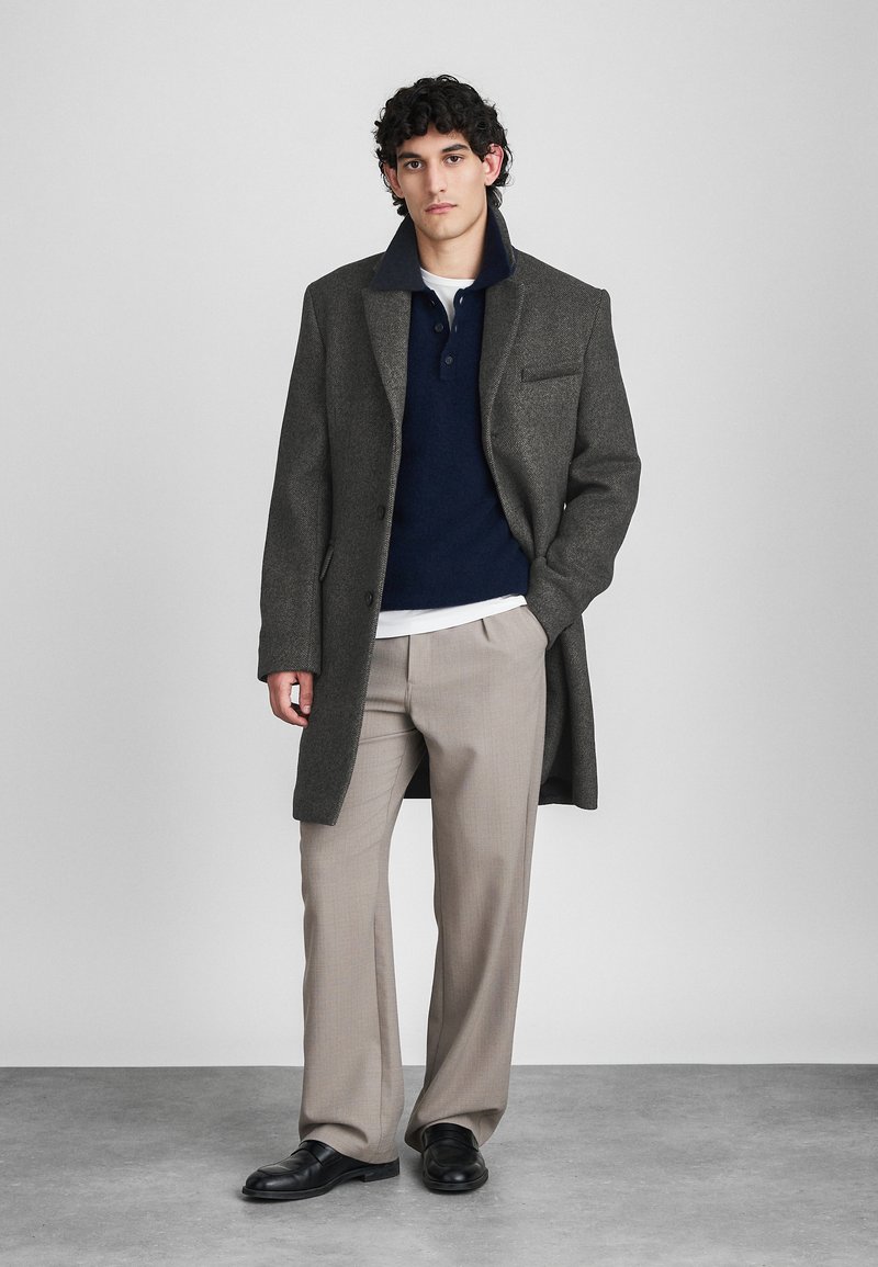 Homme se tenant dans un manteau gris, un pull bleu marine, un pantalon beige et des chaussures noires devant un fond gris clair uni.