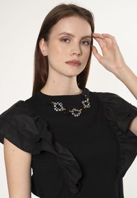 Blusa negra con mangas acampanadas, que presenta un collar de eslabones a cuadros en blanco y negro con acentos dorados. Textura suave, diseño casual.