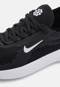 Zapatilla deportiva negra con parte superior de malla texturizada, cordones negros y un logo de Nike blanco en el lateral. Cuenta con una suela de goma blanca.