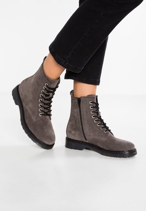 Szare, sięgające kostki zamszowe buty sznurowane, z czarnymi podeszwami i bocznymi zamkami, noszone z czarnymi krótkimi spodniami na jasnym tle.