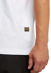 Primer plano del brazo de una persona que lleva una camisa blanca de G-Star RAW con un pequeño logo negro y amarillo cerca del dobladillo, combinada con pantalones negros.