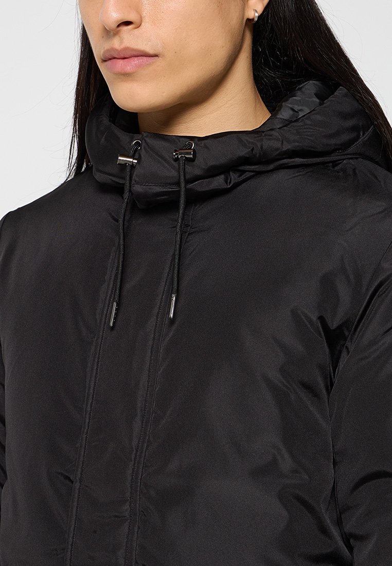 Veste matelassée noire avec un col montant, une capuche à cordon de serrage et des finitions en métal argenté. Texture lisse et design épuré et minimaliste.