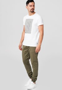 White short-sleeve t-shirt with black graphic design, paired with olive green jogger pants and white sneakers.