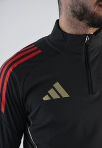 Top deportivo negro con cuello alto, que presenta franjas rojas en los hombros y un logotipo dorado de Adidas en el pecho. Cierre frontal con cremallera.