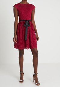 Robe en dentelle rouge avec des manches courtes, un corsage ajusté, une jupe évasée et une ceinture en satin noir à la taille. Comprend un ourlet festonné.