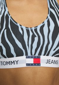 Zebramönstrad sportbh i ljus blå och svart, med ett vitt elastiskt band med "TOMMY JEANS"-logotyp och röd accent. Mjukt, elastiskt tyg.