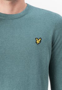 Žalias megztinis su apvalia iškirpte; krūtinėje yra maža geltona siuvinėta erelio logotipas. Minkštas pabirumas, prigludusi forma.